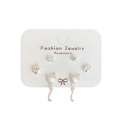 un lot de 3 paires de boucles d'oreilles argentées sur le thème des chats : deux paires de puces en forme de tête avec un strass et de patte de chat. Une paire de boucles d'oreilles en forme de tête de chat d'où pend une adorable queue de chat. L'ensemble est dans un style minimaliste et adorable.