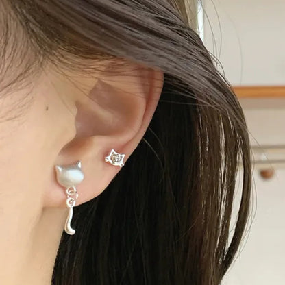 un lot de 3 paires de boucles d'oreilles argentées sur le thème des chats : deux paires de puces en forme de tête avec un strass et de patte de chat. Une paire de boucles d'oreilles en forme de tête de chat d'où pend une adorable queue de chat. L'ensemble est dans un style minimaliste et adorable.