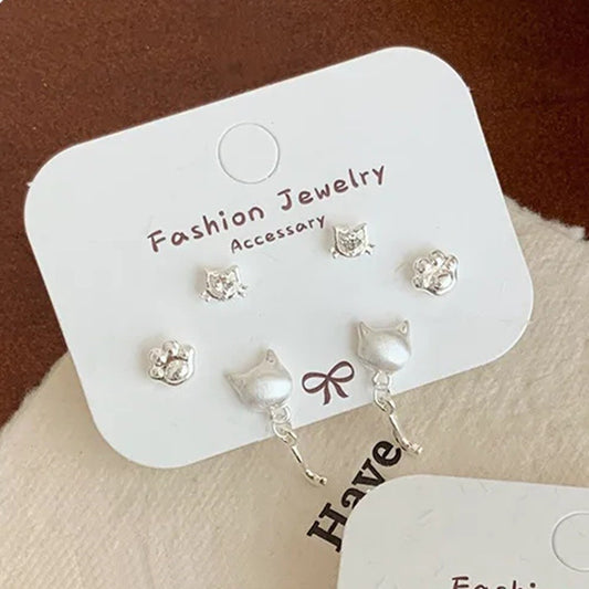 un lot de 3 paires de boucles d'oreilles argentées sur le thème des chats : deux paires de puces en forme de tête avec un strass et de patte de chat. Une paire de boucles d'oreilles en forme de tête de chat d'où pend une adorable queue de chat. L'ensemble est dans un style minimaliste et adorable.