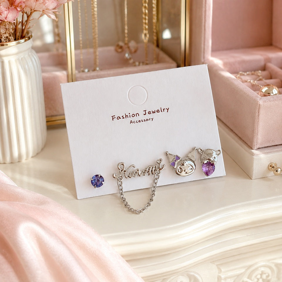 Boucles d'oreilles asymétriques Kuromi argentées avec strass violets et chaîne décorative, présentées sur un élégant dressing rose. Un bijou kawaii inspiré de l'univers japonais, idéal pour compléter une tenue ou offrir en cadeau.