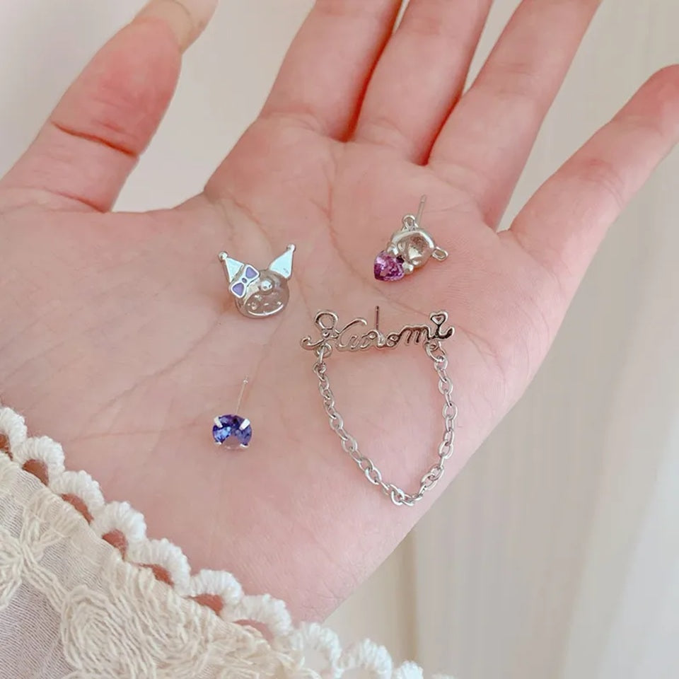 Boucles d'oreilles asymétriques Kuromi argentées avec strass violets et chaîne décorative, posées dans une main de femme. Un bijou kawaii inspiré de l'univers japonais, idéal pour compléter une tenue ou offrir en cadeau.