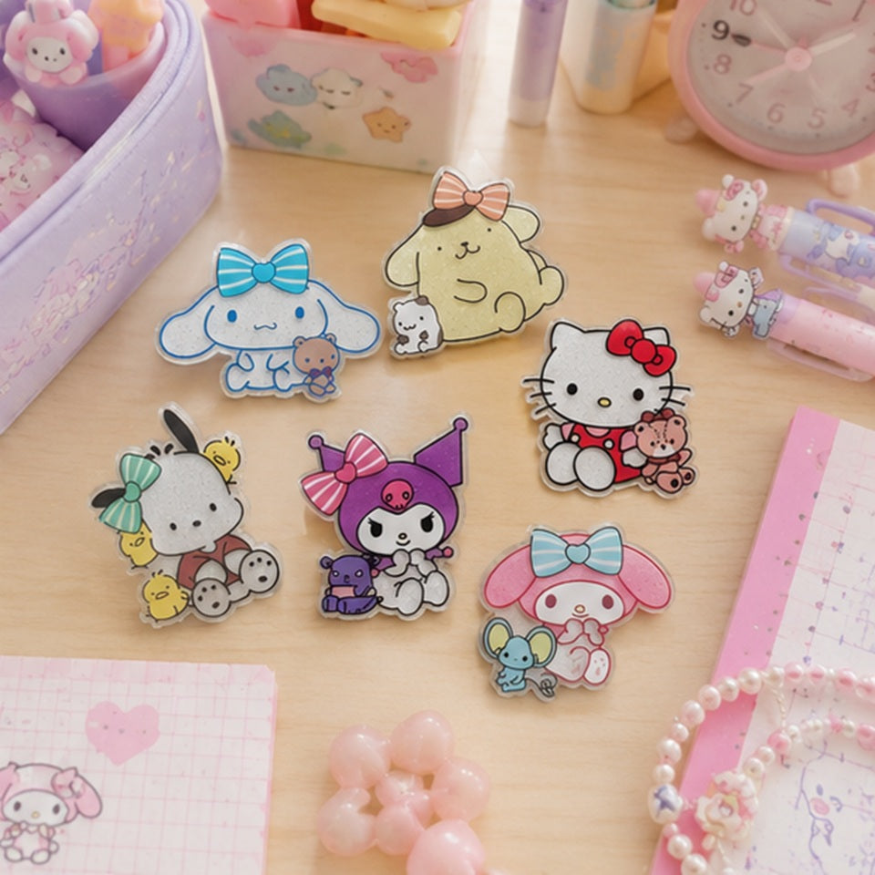 Magnets pailletés kawaii représentant plusieurs personnages Sanrio : Kuromi, cinnamoroll, Pompompurin, Hello Kitty, Pochacco et My Melody, placés sur un bureau d'écolière kawaii aux tons pastel, avec stylos roses. Ambiance douce, chaleureuse et féminine.