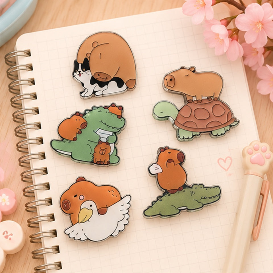 Des magnets kawaii représentant un capybara, un crocodile, un chat et un goéland sont posés sur un carnet de notes ouvert, sur un bureau mignon. Ambiance douce, dans un décor rose pastel mignon.