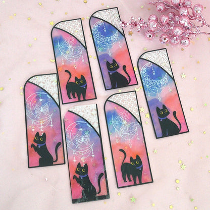des marque-pages façon vitrail coloré orné de papillons ou de lunes. Un adorable chat noir se trouve devant. L'ensemble est mystérieux et magique.