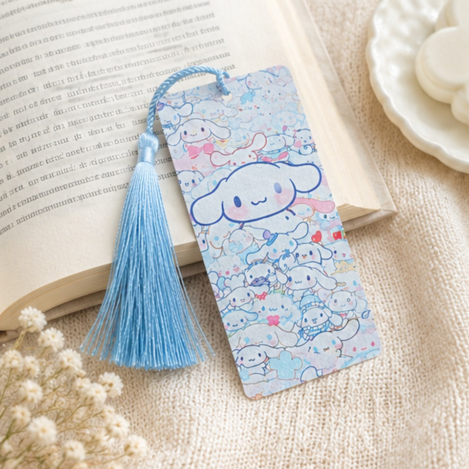Marque-page kawaii Cinnamoroll bleu avec pompon assorti, présenté sur un livre ouvert dans une ambiance douce et cosy. Accessoire de lecture Sanrio mignon aux couleurs pastel.