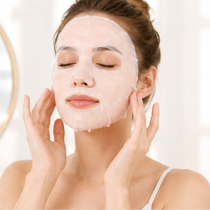 Femme de type occidental appliquant un masque en tissu hydratant à la pêche pour le visage dans une ambiance spa lumineuse : elle porte le masque sur son visage les yeux fermés. Décor doux et épuré aux tons blancs et pastel, évoquant un moment de soin et de détente.