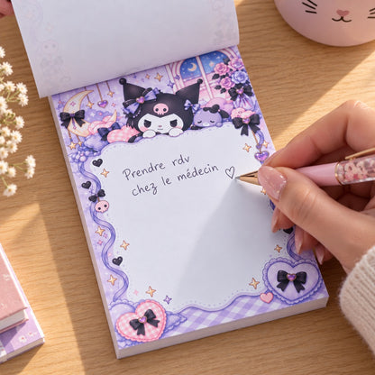 Bloc mémo rectangulaire Kuromi kawaii violet et rose pastel, décoré de cœurs, nœuds et étoiles, sur lequel une femme écrit « Prendre rdv chez le médecin » avec un stylo rose, posé sur un bureau en bois clair.