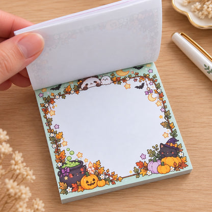 Bloc mémo carré kawaii Halloween aux couleurs pastel, décoré de chats sorciers, citrouilles, fantôme, chaudron magique et feuilles d’automne. Une main soulève les feuilles pour dévoiler une grande zone d’écriture blanche.