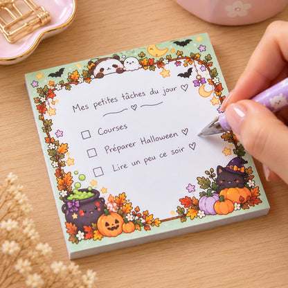 Bloc mémo carré kawaii Halloween décoré de chats sorciers, citrouilles, chauves-souris et feuilles d’automne. Une main féminine écrit une liste de tâches en français avec un stylo violet sur ce bloc-notes aux couleurs pastel.