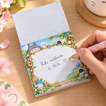 Main écrivant « Rdv médecin à 16h » sur un bloc mémo kawaii Totoro, décoré de fleurs de sakura et de personnages du Studio Ghibli, posé sur un bureau en bois avec un stylo rose.