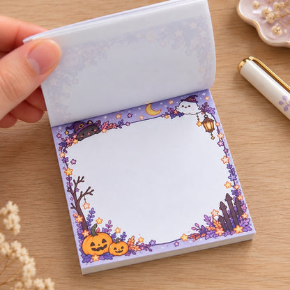 Bloc mémo kawaii Halloween aux couleurs pastel, décoré d’un chat sorcier, d’un petit fantôme, de citrouilles et d’étoiles. Une main soulève les feuilles pour dévoiler l’espace d’écriture blanc.
