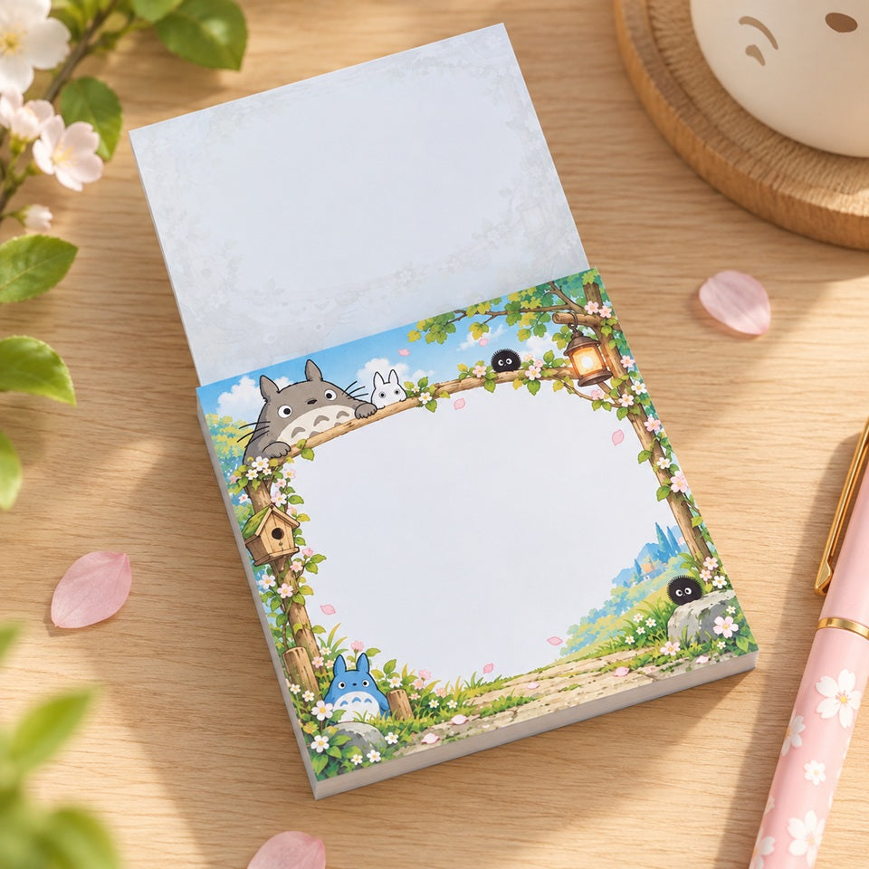 Bloc mémo kawaii Totoro illustré de fleurs de sakura, de verdure et de petits personnages Ghibli, avec une grande zone blanche pour écrire, présenté sur un bureau en bois avec un stylo rose.
