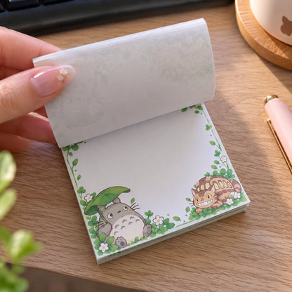Bloc mémo carré kawaii Totoro, décoré de fleurs et feuillages verts, avec Totoro et le Chatbus. Une main féminine soulève les feuilles du mémo posé sur un bureau en bois clair, dévoilant un grand espace blanc pour écrire ses notes et petits messages