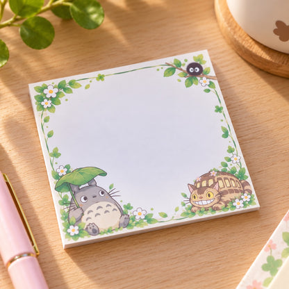 Bloc mémo carré kawaii Totoro, décoré de feuillages verts, de petites fleurs blanches, de Totoro sous sa feuille, du Chatbus et d'une Noiraude. Posé sur un bureau en bois clair, ce mémo au style Studio Ghibli offre un grand espace blanc pour écrire ses notes et petits messages.