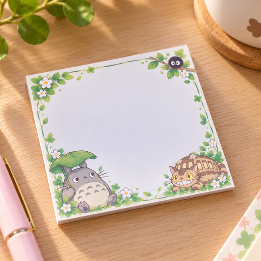 Bloc mémo carré kawaii Totoro, décoré de feuillages verts, de petites fleurs blanches, de Totoro sous sa feuille, du Chatbus et d'une Noiraude. Posé sur un bureau en bois clair, ce mémo au style Studio Ghibli offre un grand espace blanc pour écrire ses notes et petits messages.