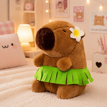 Peluche capybara marron toute douce portant une jupe hawaïenne verte et une fleur blanche à l'oreille, posée sur un lit dans une jolie chambre aux tons pastel. En arrière-plan, on aperçoit un coussin à motif floral, une lampe allumée, un vase de roses et de petites décorations kawaii créant une ambiance chaleureuse et mignonne.