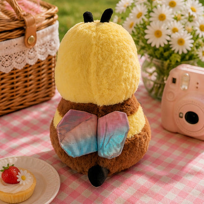 Peluche abeille kawaii vue de dos avec ailes colorées irisées, dans un décor pique-nique rose avec marguerites, panier en osier et appareil photo pastel.