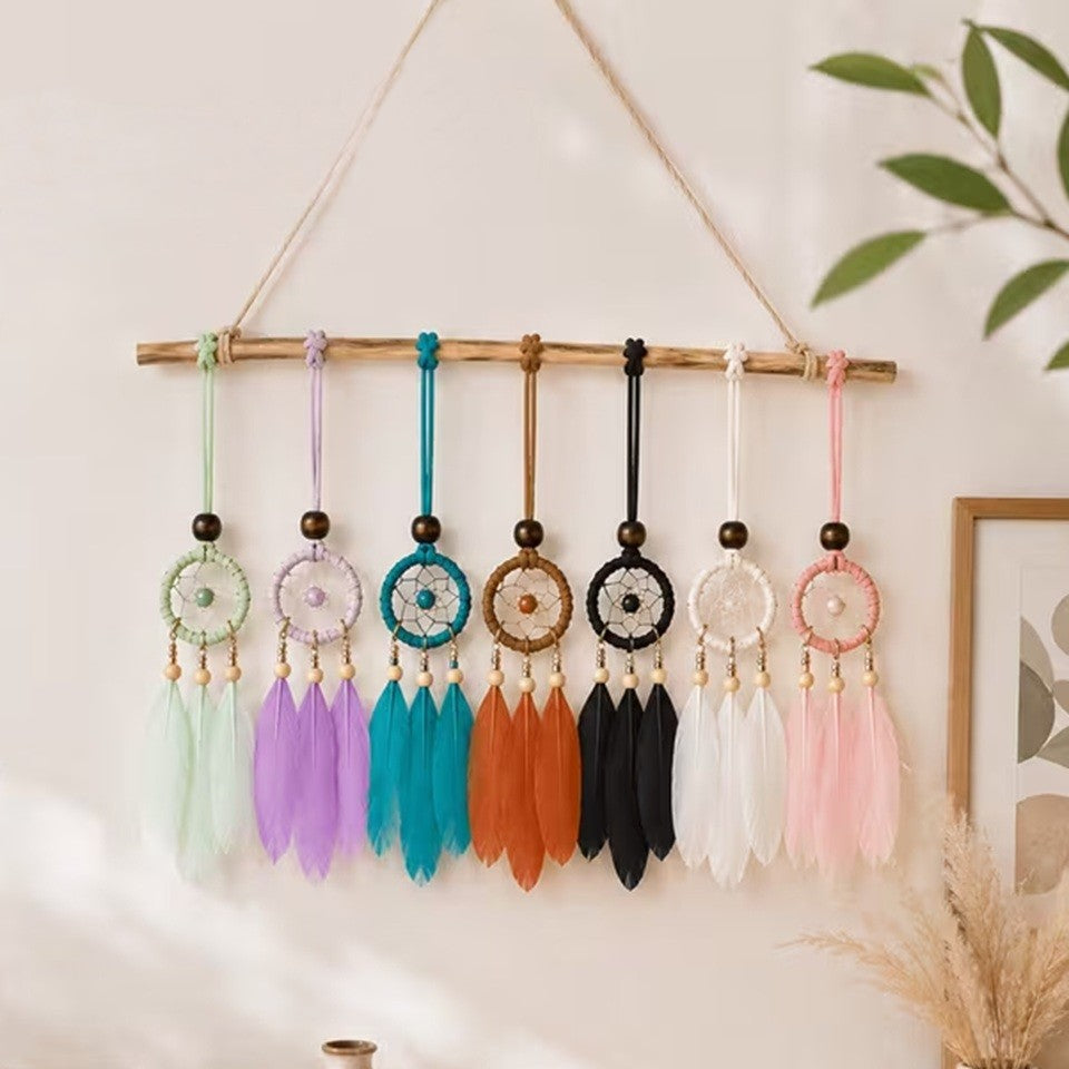 des suspensions attrape-rêves miniatures avec trois plumes, déclinées en plusieurs coloris : vert menthe, rose pastel, bleu turquoise, blanc, noir et orange. Les suspensions sont mises en scène comme décoration murale dans une ambiance douce et chaleureuse. Chaque modèle est orné d'un cercle tissé, de perles en bois et de plumes colorées, avec un style bohème et artisanal.