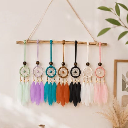 des suspensions attrape-rêves miniatures avec trois plumes, déclinées en plusieurs coloris : vert menthe, rose pastel, bleu turquoise, blanc, noir et orange. Les suspensions sont mises en scène comme décoration murale dans une ambiance douce et chaleureuse. Chaque modèle est orné d'un cercle tissé, de perles en bois et de plumes colorées, avec un style bohème et artisanal.