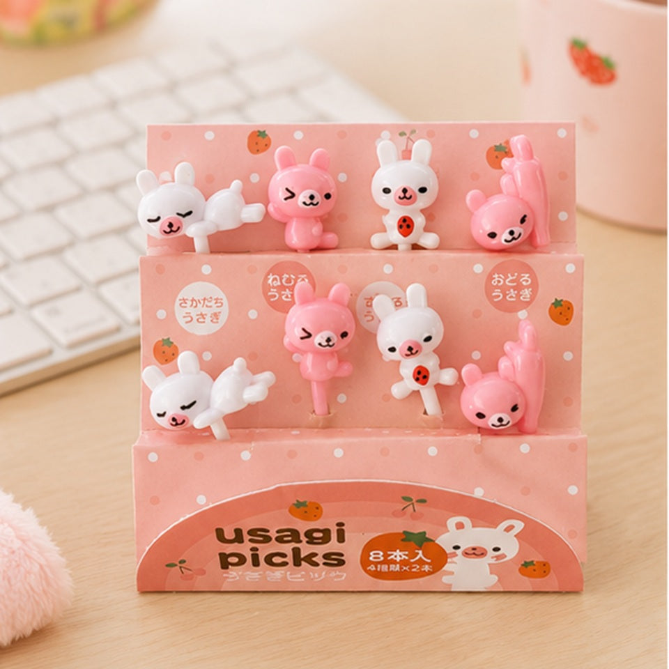 Lot de pics alimentaires lapins kawaii roses et blancs dans leur présentoir Usagi Picks, mis en scène dans un décor pastel avec figurine lapin.