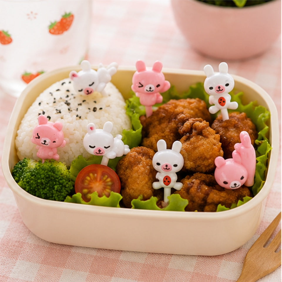 Pics décoratifs kawaii en forme de lapins roses et blancs utilisés dans une boîte bento avec riz, poulet et légumes.