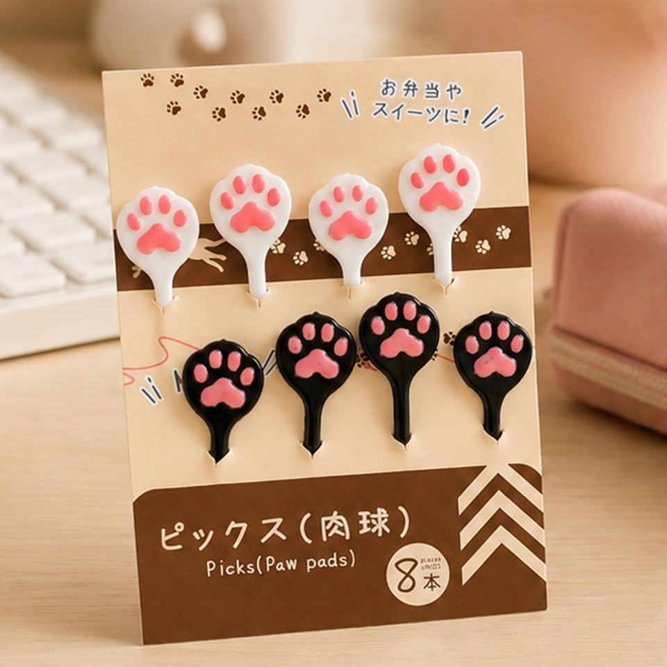 Pics alimentaires kawaii en forme de pattes de chat roses, noirs et blancs, présentés dans leur emballage sur un bureau en bois.