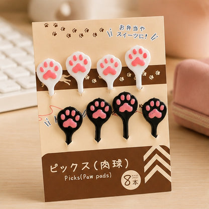 Pics alimentaires kawaii en forme de pattes de chat roses, noirs et blancs, présentés dans leur emballage sur un bureau en bois.