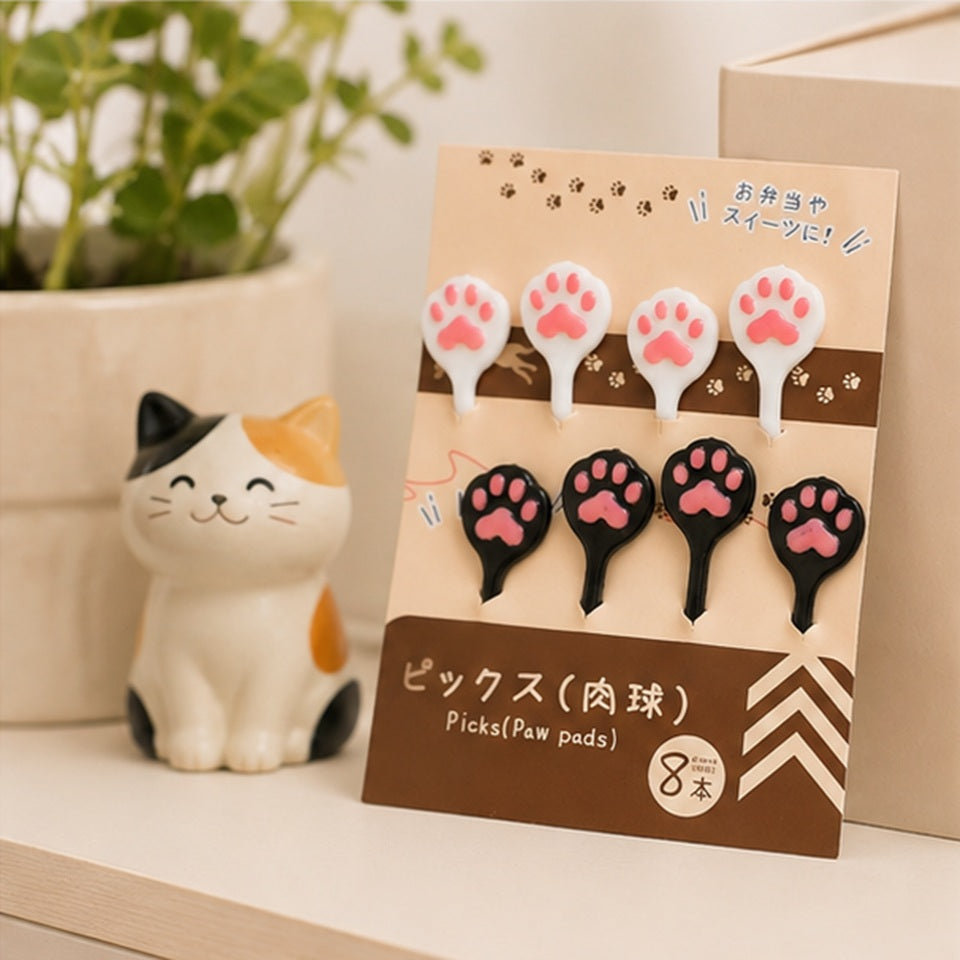 Pics kawaii en forme de pattes de chat roses, noirs et blancs dans leur emballage, présentés dans un décor doux avec figurine de chat.