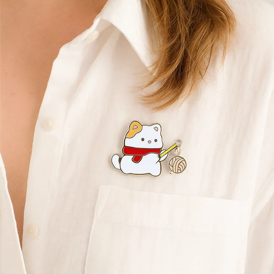 un pin's adorable représentant un chat blanc portant une écharpe qui porte un baton au bout duquel est suspendu une pelote de laine.