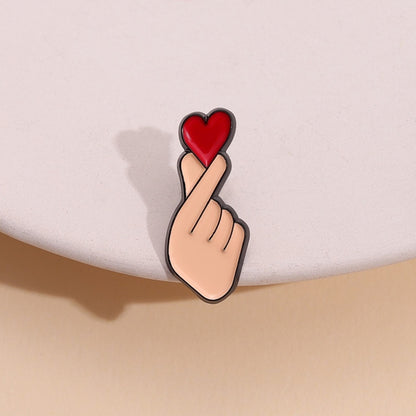 un pin's en forme de main qui forme un coeur coréen, le finger heart. Il y a un gros coeur rouge au dessus, symbole de l'amour