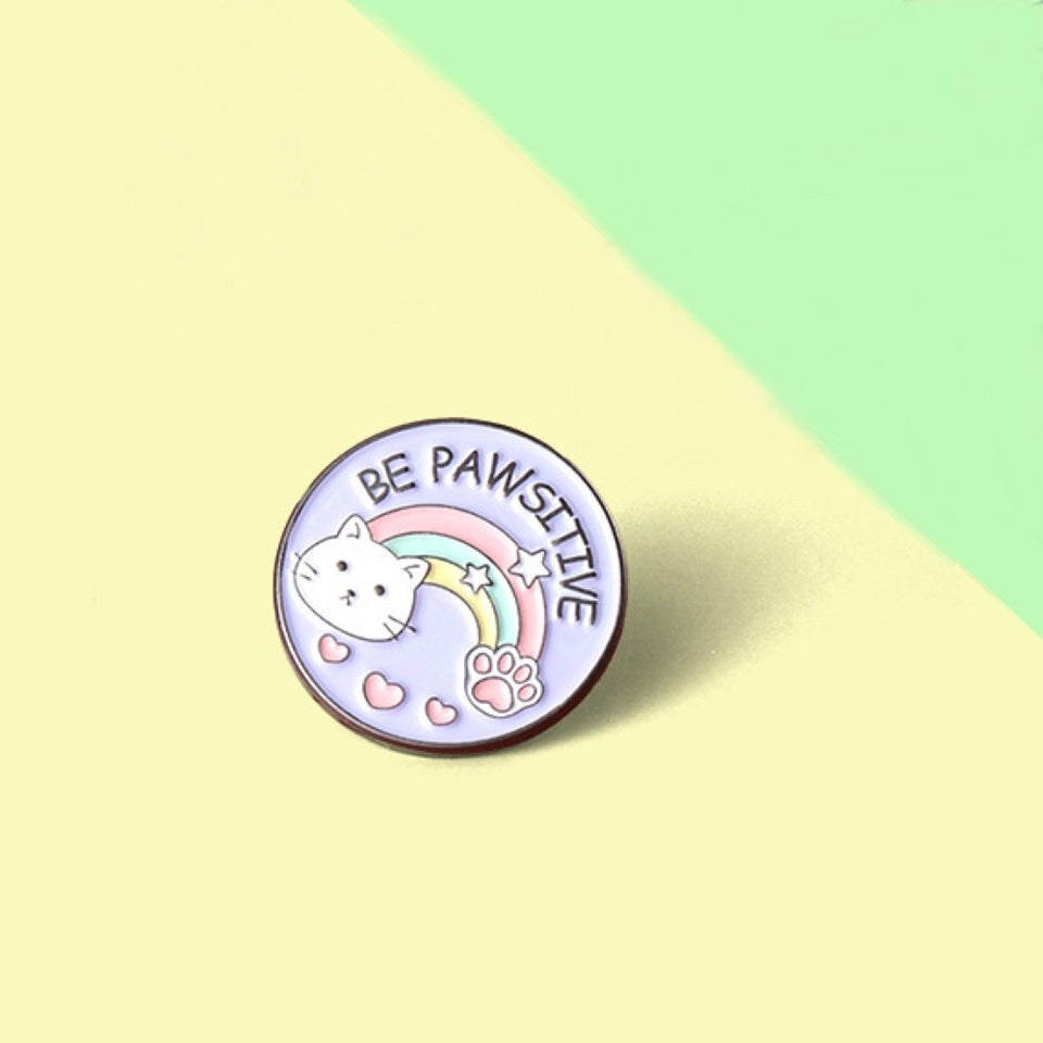 un pin's rond au design d'un adorable chat et d'un arc-en-ciel et d'une patte de chat. Le message Be Pawsitive y est inscrit. En plus du jeu de mot, il véhicule un message positif qui fait du bien au moral