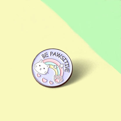 un pin's rond au design d'un adorable chat et d'un arc-en-ciel et d'une patte de chat. Le message Be Pawsitive y est inscrit. En plus du jeu de mot, il véhicule un message positif qui fait du bien au moral