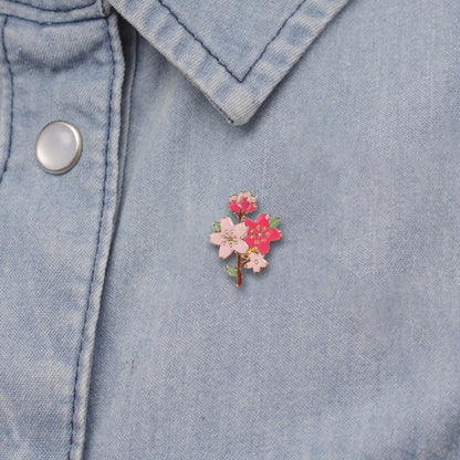 un pin's doré en forme et aux couleurs d'une branche de cerisier en fleurs. On y trouve des jolies fleurs de sakura rose foncé et rose clair et des boutons de fleurs. L'ensemble est poétique