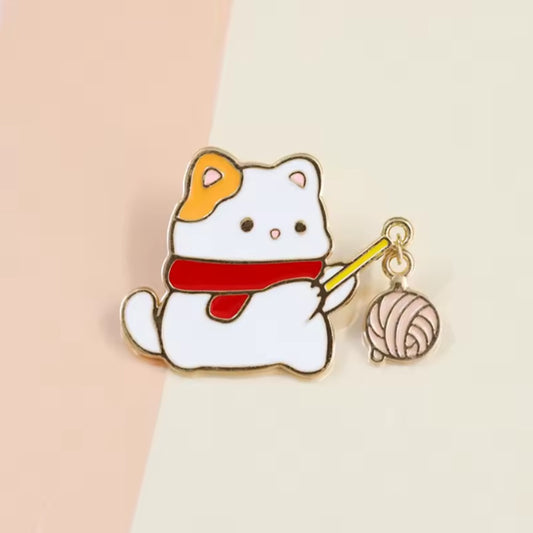 un pin's adorable représentant un chat blanc portant une écharpe qui porte un baton au bout duquel est suspendu une pelote de laine.