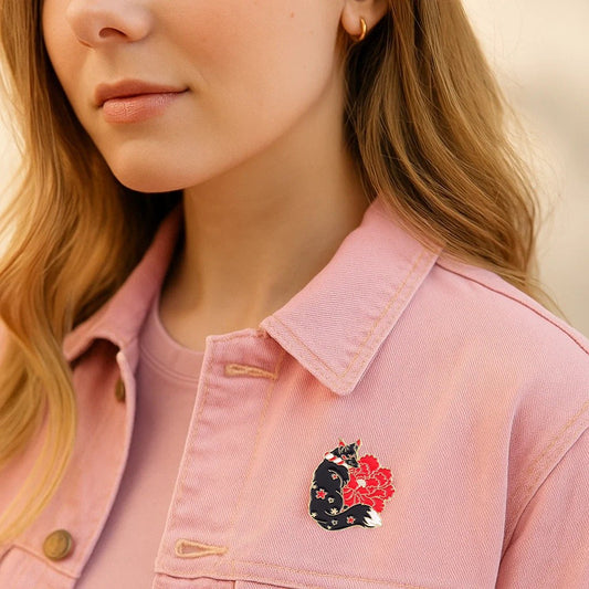 un pin's en forme de kitsune noir dont la fourrure est ornée de fleurs de sakura rouges. Une grosse fleur rouge se trouve derrière le renard. L'ensemble est poétique, rempli de symbole