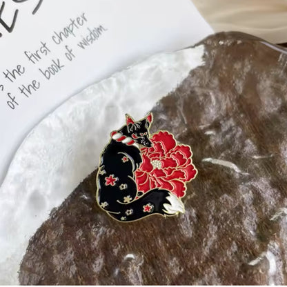 un pin's en forme de kitsune noir dont la fourrure est ornée de fleurs de sakura rouges. Une grosse fleur rouge se trouve derrière le renard. L'ensemble est poétique, rempli de symbole