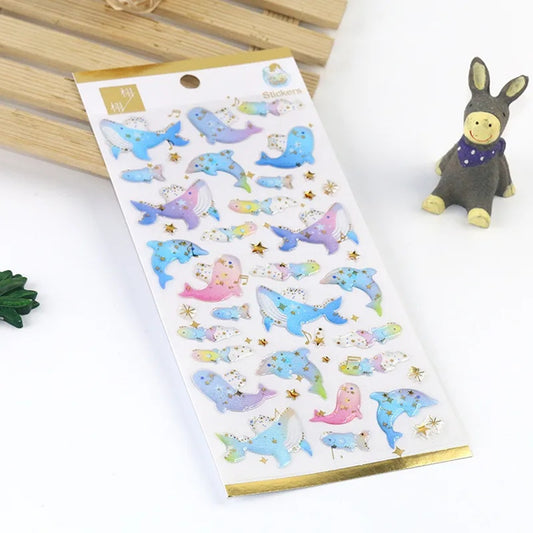 une planche de stickers en forme de baleine et de dauphin, dans les tons de couleurs bleu et violet. Ils ont des étoiles dorés qui apportent de la brillance. Effet poétique et mystérieux