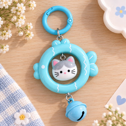 Porte-clés kawaii bleu en forme de poisson avec petit chat gris et blanc et grelot assorti, posé à plat dans un décor doux et fleuri.