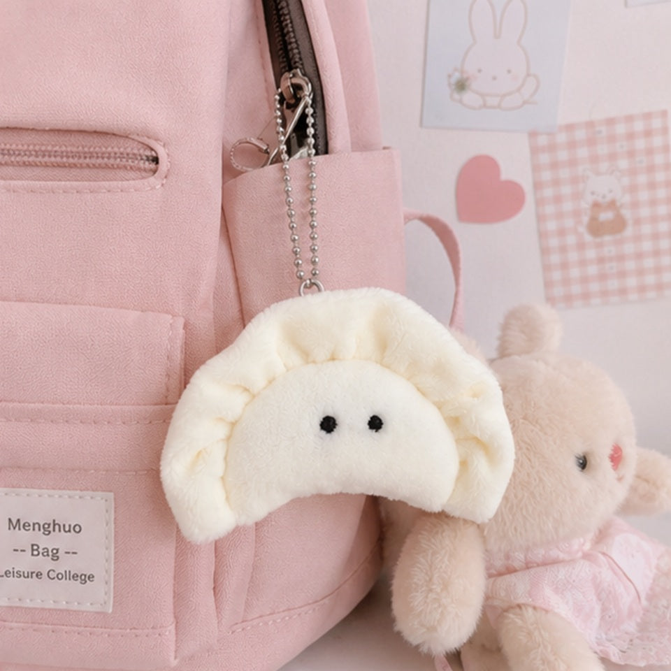 un porte-clés peluche en forme de gyoza blanc crème avec de petits yeux noirs brodé suspendu à un sac rose grâce à une chaîne argentée à billes. Ambiance chaleureuse et mignonne, féminine et kawaii.