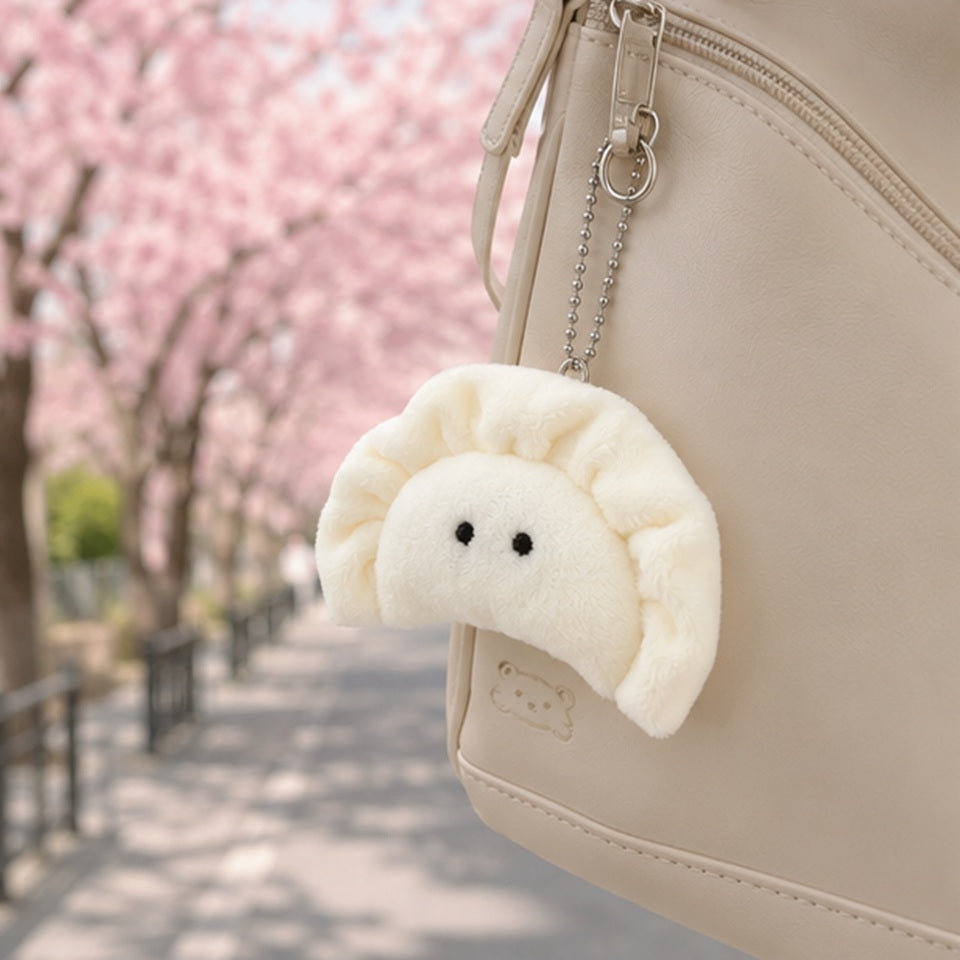 un porte-clés peluche en forme de gyoza blanc crème avec de petits yeux noirs brodé suspendu à un sac grâce à une chaîne argentée à billes. Dans un style kawaii avec des fleurs et des couleurs douces créant une ambiance chaleureuse et mignonne.