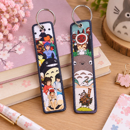 Deux marque-pages porte-clés inspirés de l’univers Studio Ghibli, avec Totoro, Chihiro et Princesse Mononoké, posés sur un bureau en bois avec des livres roses à fleurs de sakura et des accessoires kawaii.