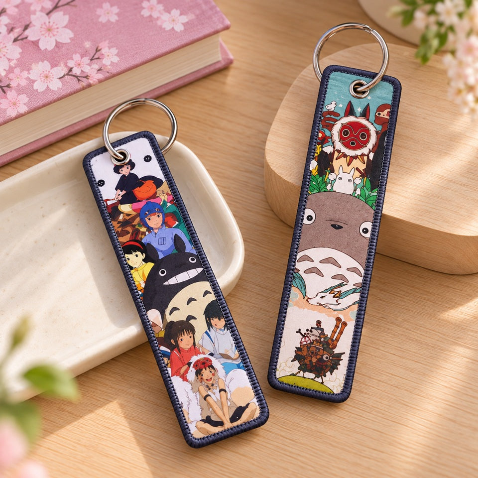 Deux grands porte-clés rectangulaires Studio Ghibli avec Totoro, Chihiro, Kiki et Princesse Mononoké, posés sur un plateau en céramique et un support en bois, près d’un carnet rose à fleurs de sakura.