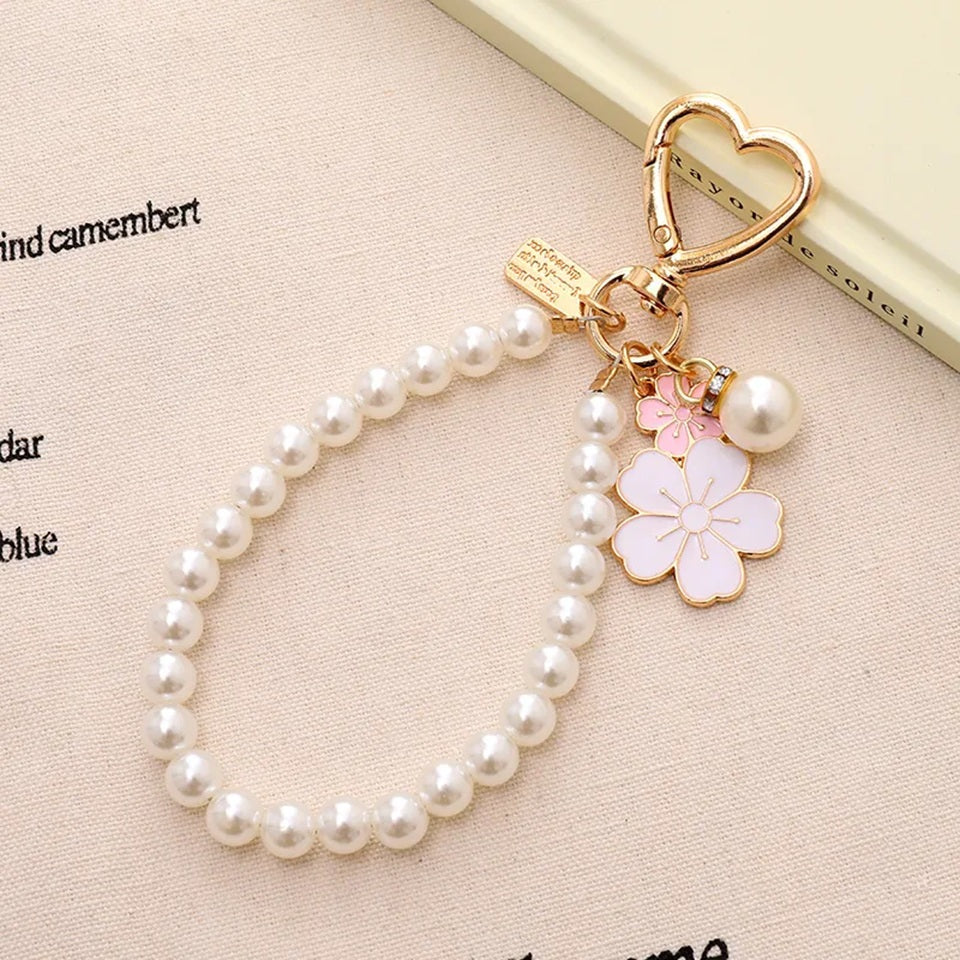 Porte-clé bijou avec perles blanches, mousqueton cœur doré et pendentifs fleurs de sakura roses, présenté dans un décor rose pastel élégant.