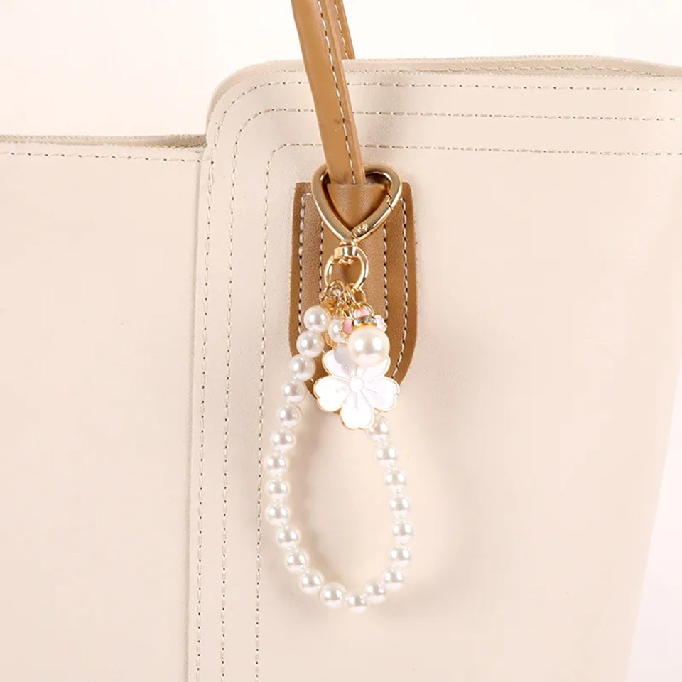 Porte-clé bijou avec perles blanches, mousqueton cœur doré et pendentifs fleurs de sakura roses accroché sur une anse d'un sac à main.
