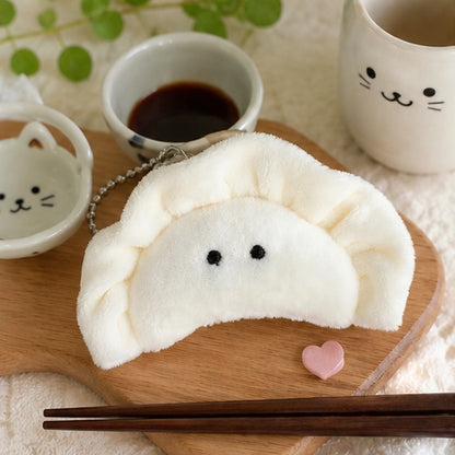 Porte-clés peluche kawaii en forme de gyoza  mignon blanc crème est présenté sur une planche en bois marron, accompagné d'une tasse de café et divers éléments de vaisselle kawaii inspirés de l'univers kawaii japonais. Idéal pour personnaliser un sac, un sac à dos ou un trousseau de clés.
