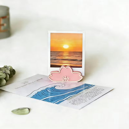 un porte-photo en forme de fleur sakura rose dans lequel est posé une photo d'un coucher de soleil. L'ambiance est douce et calme. Idéal pour une touche décoration florale mignonne