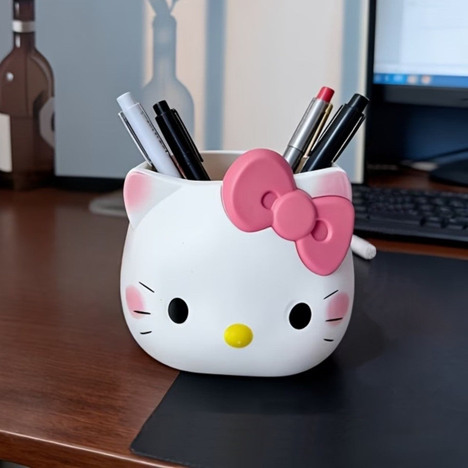 un pot à crayon blanc Hello Kitty avec son noeud rose posé sur un bureau en chêne brun sur lequel se trouve un ordinateur. Ambiance studieuse mais détail mignon et coloré !