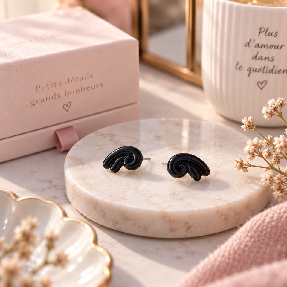 Puces d’oreilles noires au design tourbillon, présentées sur un support effet pierre dans un décor élégant, féminin et romantique.
