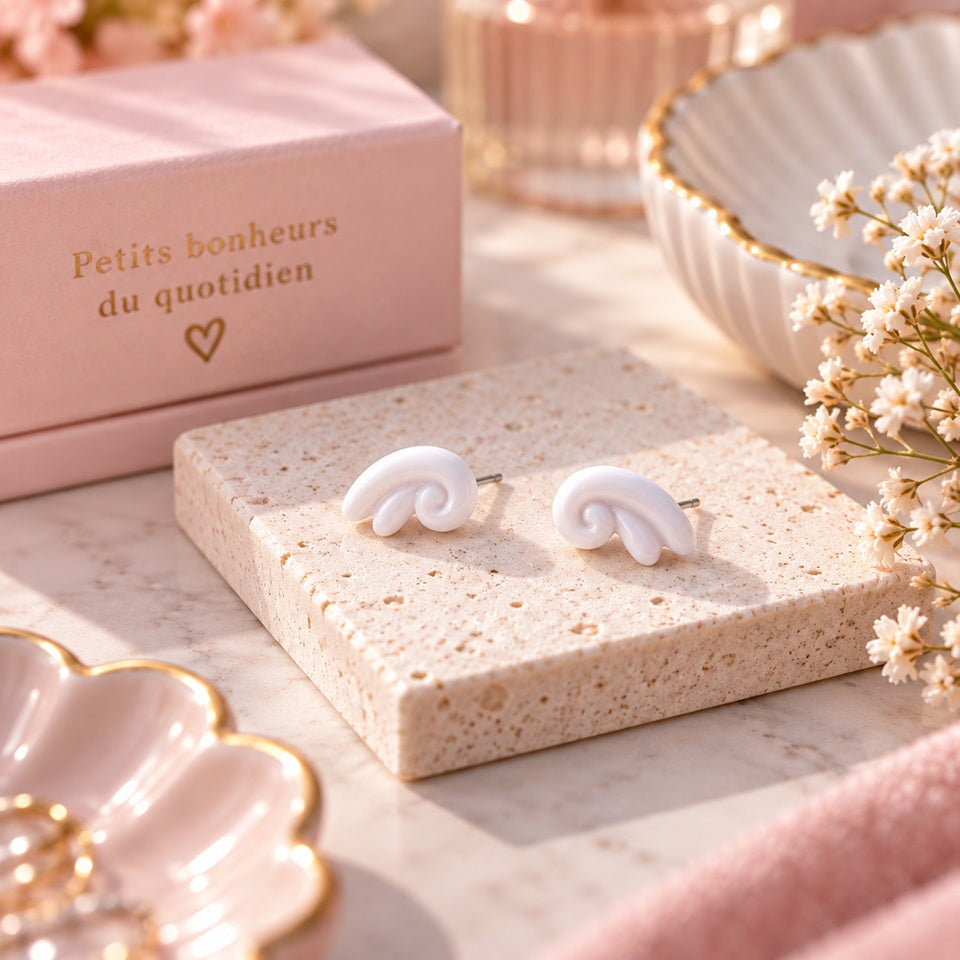 Puces d’oreilles blanches au design tourbillon, présentées sur un support en pierre dans un décor romantique rose et fleuri.