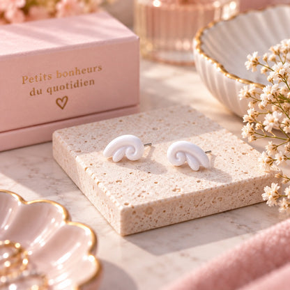 Puces d’oreilles blanches au design tourbillon, présentées sur un support en pierre dans un décor romantique rose et fleuri.
